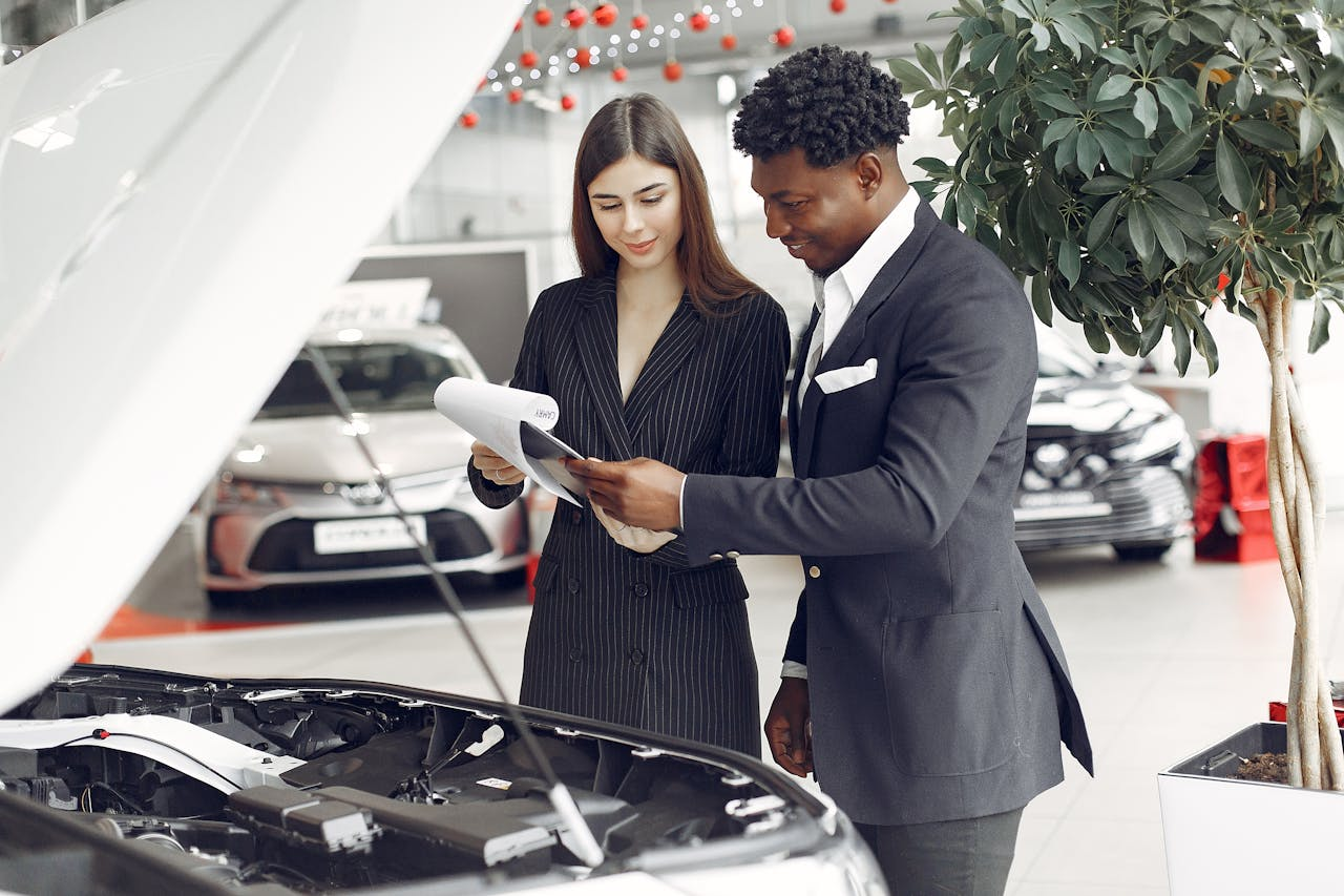 digital Man and woman reviewing documents at a car dealership next to an open car hood.